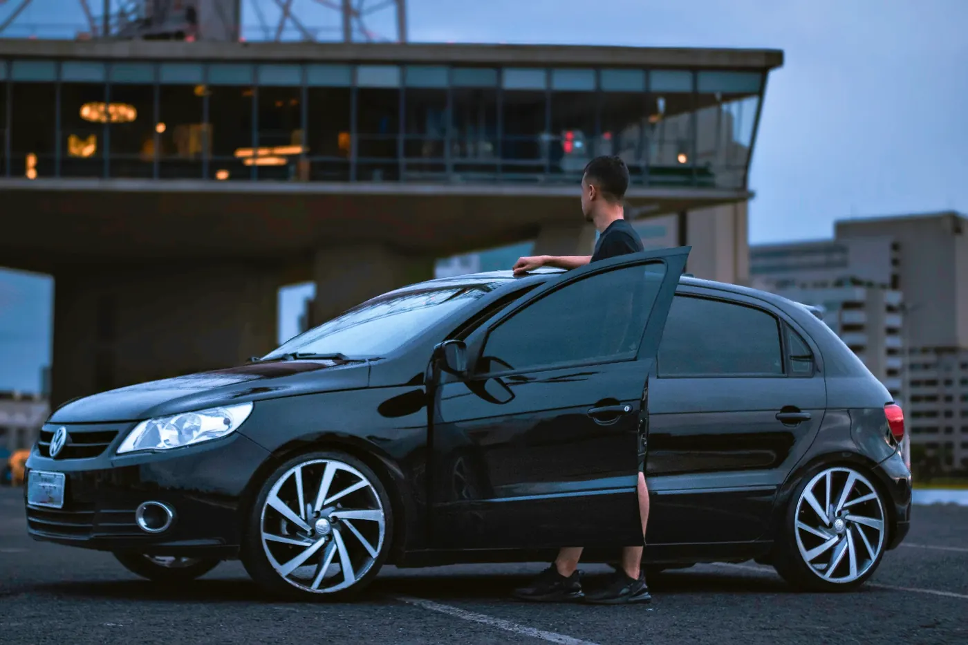 Homem ao lado de um carro preto de porta aberta, ao anoitecer