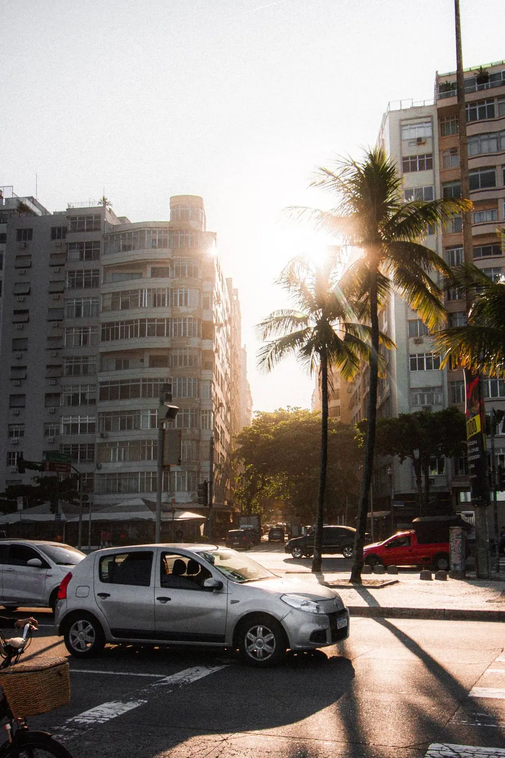 Carro prata em um cruzamento com palmeiras e prédios, na luz do fim de tarde